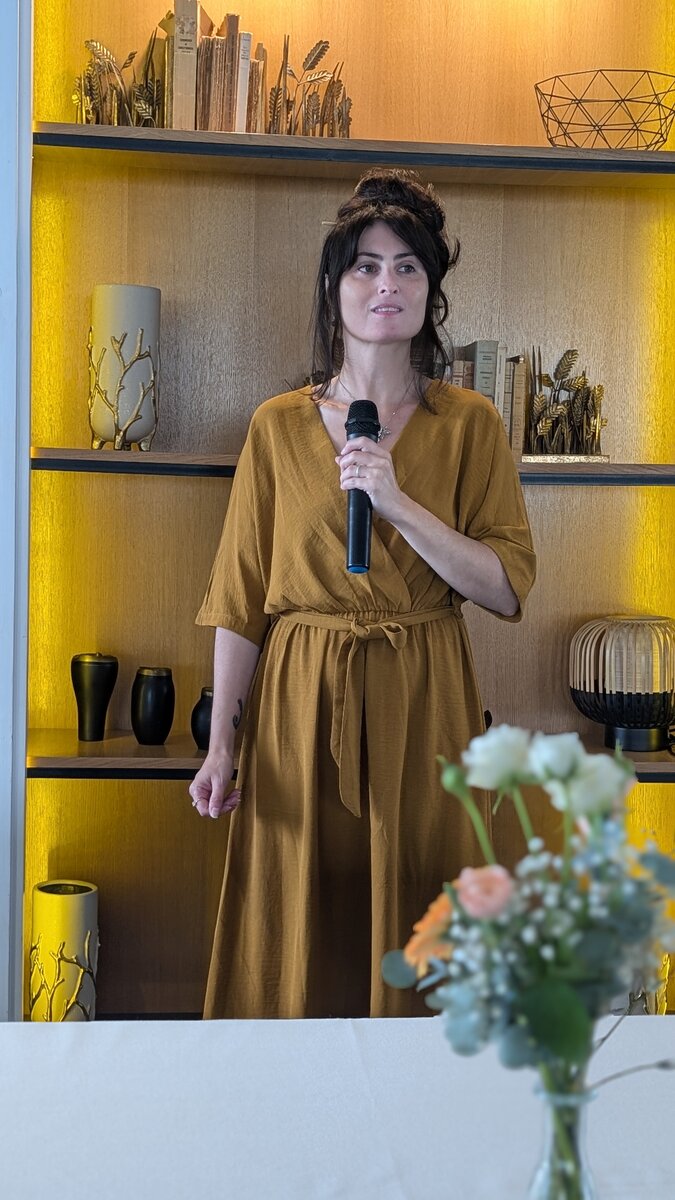Mylène Chevreul debout avec un micro devant une bibliothèque éclairée, près d'un bouquet de fleurs