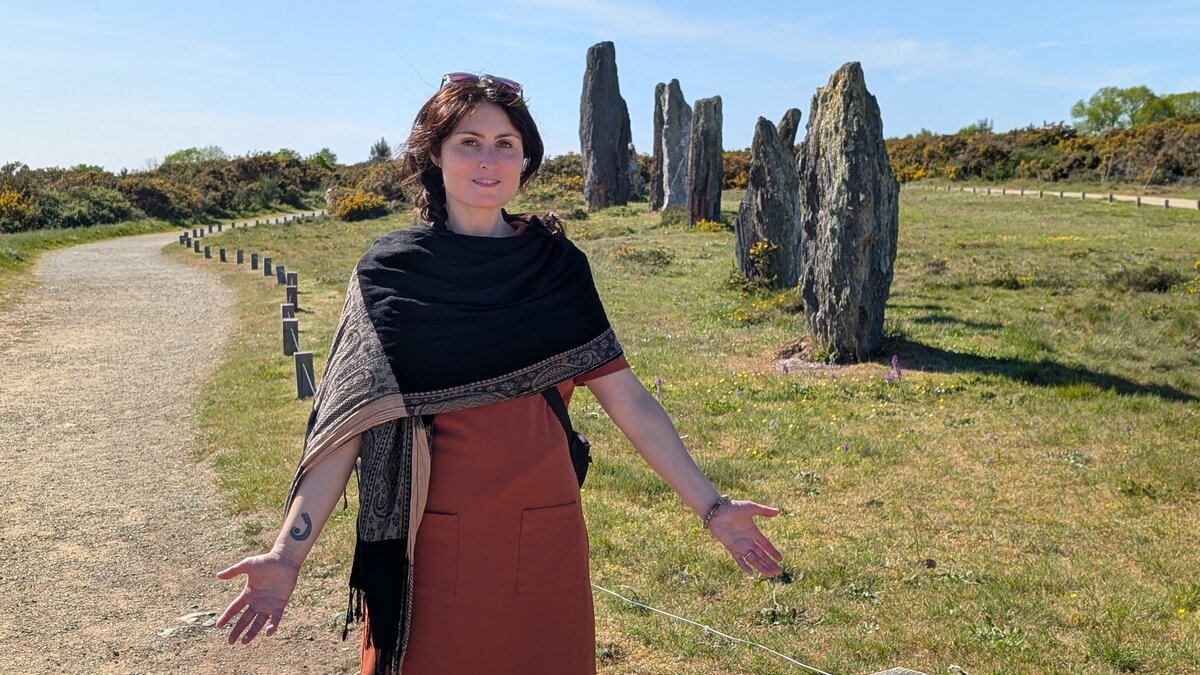 Mylène Chevreul debout devant les alignements de menhirs de Saint-Just en Bretagne, vêtue d'un châle et d'une robe terra cotta, bras ouverts vers le paysage